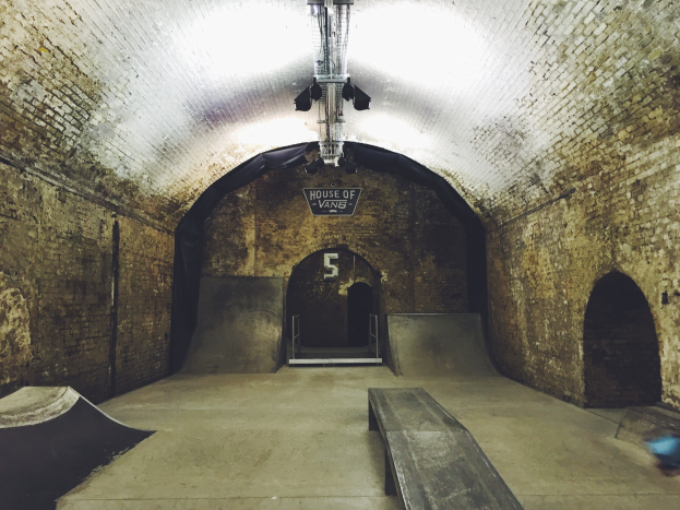 Ein Skatepark mit einer Ziegelwand-Tunnelrampe und einer Bank in der Mitte, einem Textschild und einer Deckenbeleuchtung.