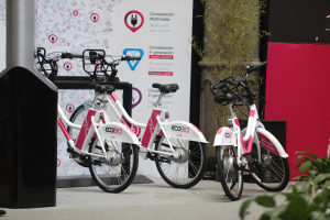 Zwei pinke und weiße Fahrräder nebeneinander vor einer roten Wand geparkt, mit einer Säule links und Pflanzen rechts und einem Banner mit Text und Logos im Hintergrund.