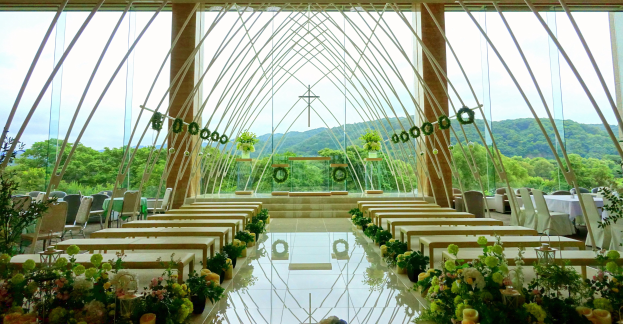 Eine Hochzeitszeremonie in einem großen Saal mit Tischen und Stühlen in einem Halbkreis angeordnet, dekoriert mit Blumenbouquets und anderen Gegenständen, vor einer Kulisse aus Bäumen, Hügeln und einem klaren blauen Himmel.