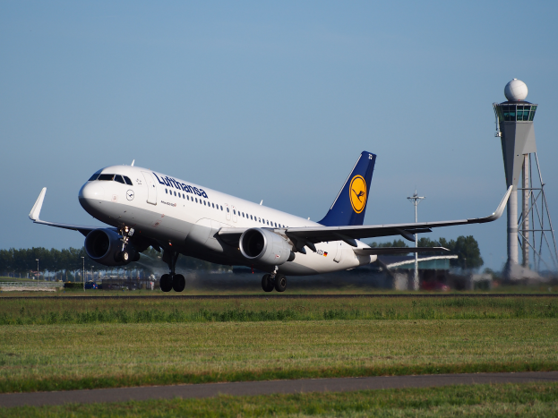 Ein Lufthansa Airbus A320-200 startet von Frankfurt Airport, mit grünem Gras und einer Straße im Vordergrund und hohen Türmen, Bäumen und einem klaren blauen Himmel im Hintergrund.