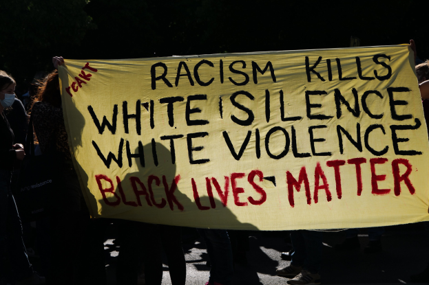 Eine Gruppe maskierter Menschen steht auf einer dunklen Straße und hält ein Banner mit der Aufschrift "Rassismus tötet Weiße Stille, weiße Gewalt, Black Lives Matter".