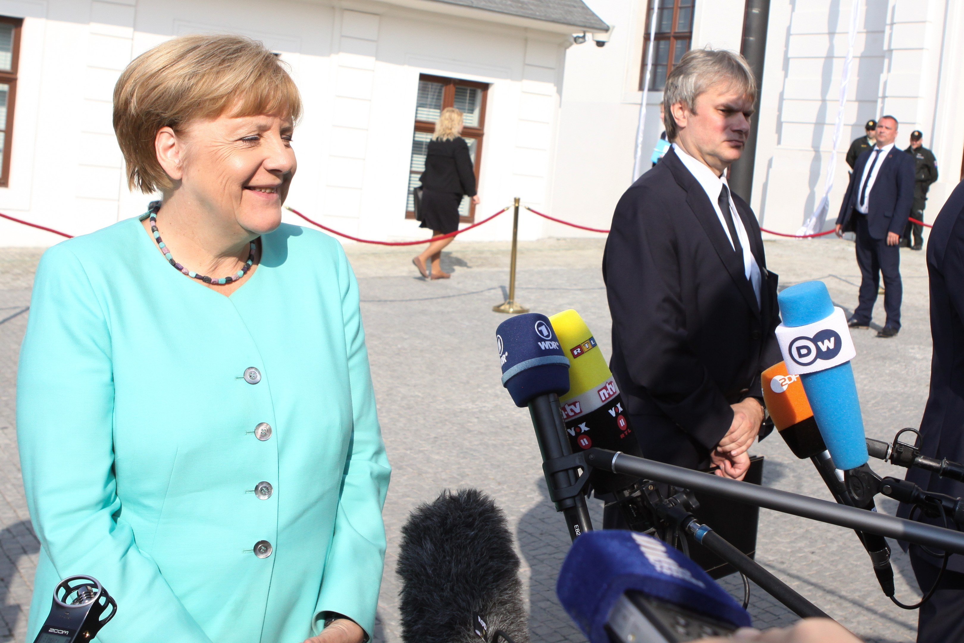 Bundeskanzlerin Angela Merkel beim Sprechen mit Reportern vor einem Gebäude mit einer Gruppe von Menschen mit Mikrofonen und einer Seilabsperrung in einer bewölkten Umgebung.