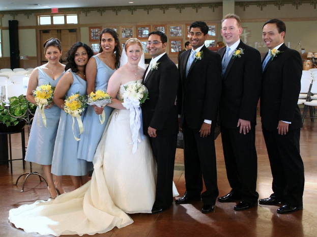 Eine Frau in einem weißen Kleid mit einem Strauß steht in der Mitte, umgeben von Frauen in blauen Kleidern mit Strauß und Männern in schwarzen Anzügen, die alle lächeln und drinnen mit verschiedenen Gegenständen und Dekorationen im Hintergrund stehen.