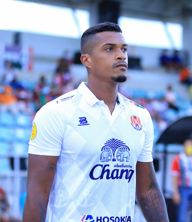 Ein Mann in einem weißen T-Shirt mit Tätowierungen auf den Händen steht auf einem Fußballfeld, mit einer unscharfen Gruppe von Menschen im Hintergrund.