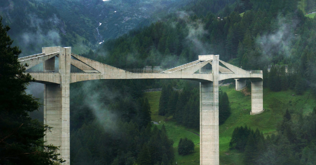 Eine Brücke auf einem Weg mit Bäumen, Hügeln und Nebel im Hintergrund.