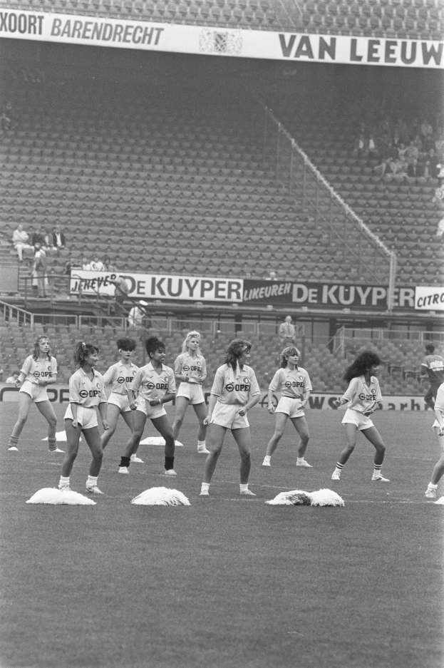 Schwarzes und weißes Bild von Frauen auf einem Fußballfeld mit Zuschauern auf den Rängen und Bannern im Hintergrund.