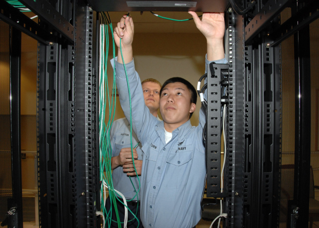 Zwei Männer arbeiten an einem Server in einem Rechenzentrum mit Regalen voller Kabel und einer Wand im Hintergrund.