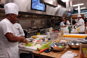 Eine Gruppe von Köchen in einer Küche, die Essen auf einem Schneidbrett zubereiten, mit einem Koch im Vordergrund in einer weißen Schürze und Mütze, der ein Messer hält und von verschiedenen Utensilien, Schüsseln, Gemüse, einem Mobiltelefon, Flaschen und anderen Gegenständen umgeben ist, mit einem Fernseher und Deckenleuchten im Hintergrund.