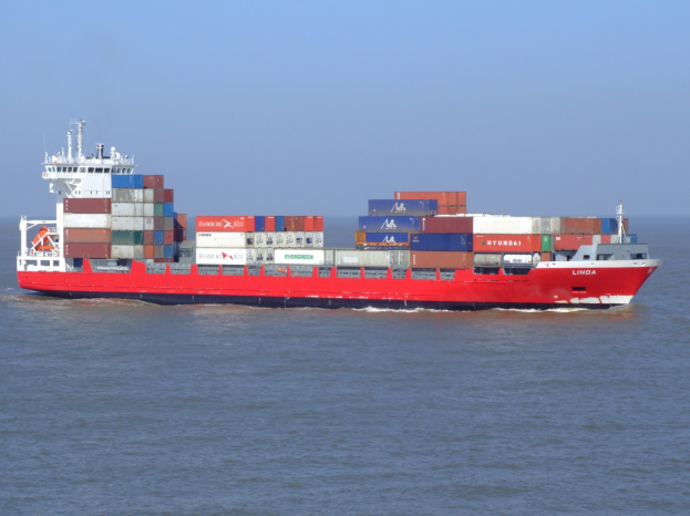 Ein großes rotes und weißes Containerschiff mit vielen Containern auf seinem Deck fährt auf dem Meer unter einem klaren blauen Himmel.