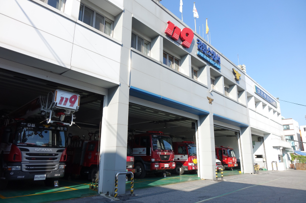 Feuerwache mit parkenden Feuerwehren, Säulen, Fenstern, Namensschildern und Flaggen im Vordergrund, vor anderen Gebäuden und einem klaren blauen Himmel.