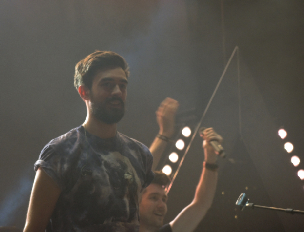 Ein Mann in einem T-Shirt steht und lacht, mit einem anderen lachenden Mann, Lichtern, Rauch, einem Mikrofon und anderer Ausr├╝stung im Hintergrund.