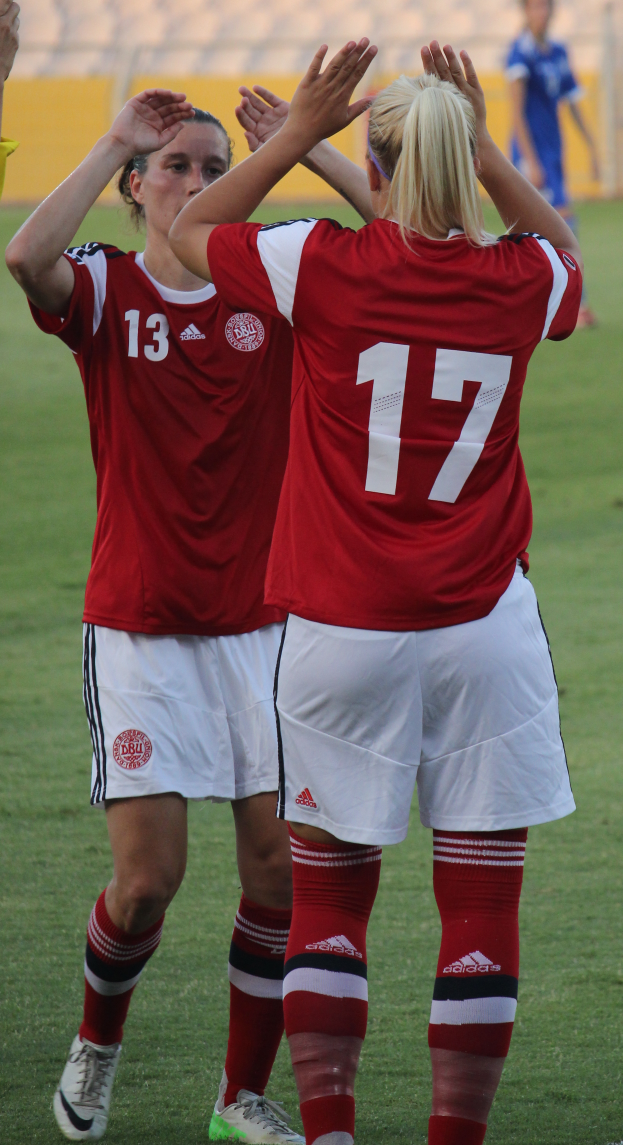 Zwei Frauen in roten T-Shirts und weißen Shorts stehen auf einem Fußballfeld, klatschen in die Hände und feiern, mit einer Person in einem blauen Kleid, einer gelben Tafel und ein paar Stühlen im Hintergrund.