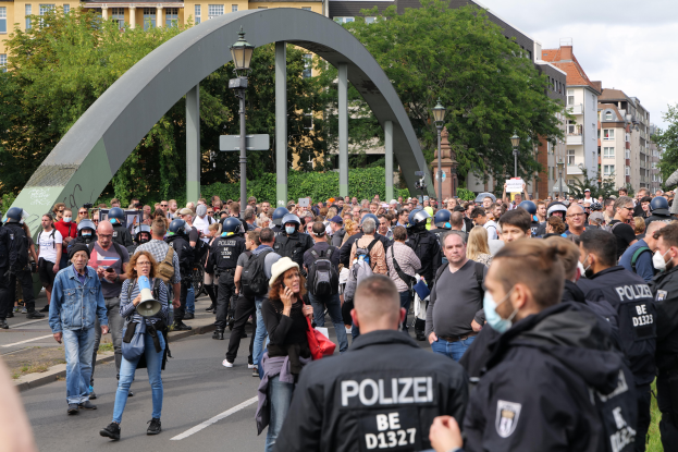 Eine große Gruppe von Menschen geht eine Straße entlang, die von Polizisten gesäumt ist, einige tragen Mützen und Masken, andere tragen Taschen, einer hält ein Megafon, Laternenpfähle, ein Tor, Bäume, Gebäude mit Fenstern und ein bewölkter Himmel im Hintergrund.