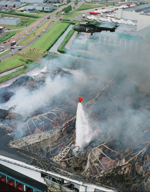Ein Hubschrauber fliegt über ein großes, durch ein Feuer zerstörtes Gebäude, aus dem Rauch aufsteigt, mit mehreren Gebäuden, Fahrzeugen und Bäumen im Hintergrund und Text unten.
