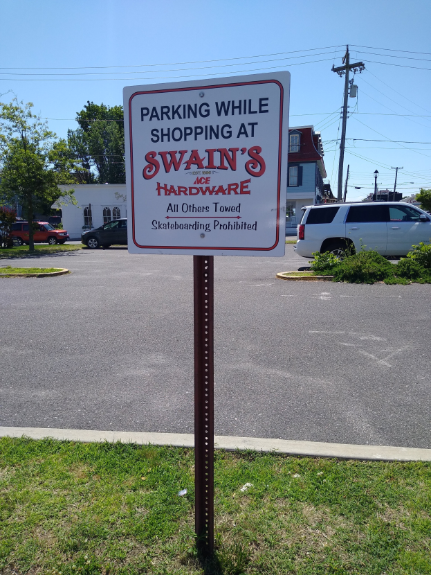 Ein Parkplatz mit einem Schild, auf dem "Parken während des Einkaufs bei Swain's Hardware" steht, im Vordergrund, mit Fahrzeugen, Pflanzen, Bäumen, Gebäuden, Strommasten mit Drähten und einem klaren blauen Himmel im Hintergrund.