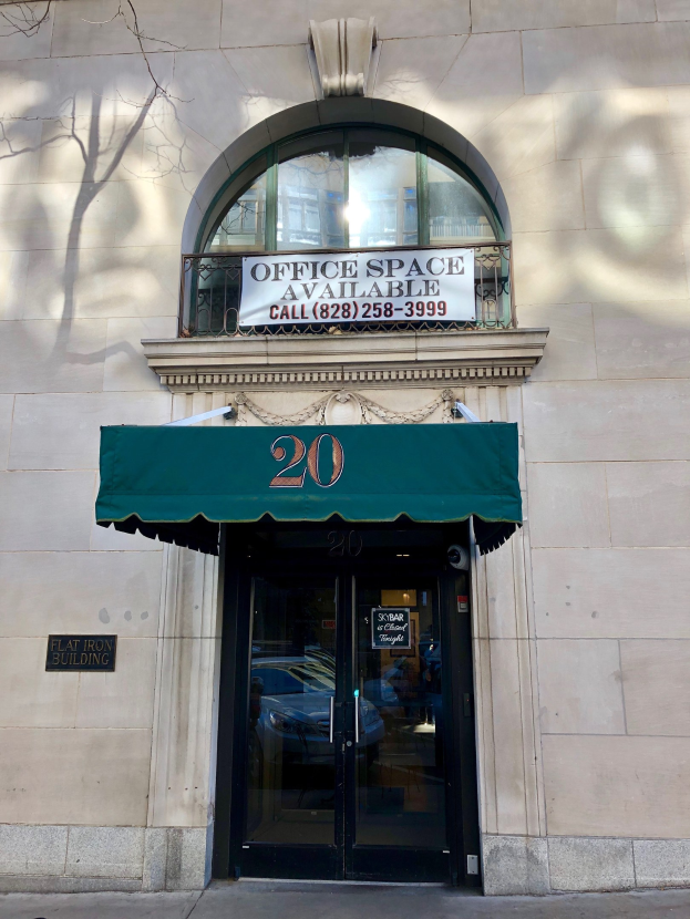 Ein Gebäude mit einer Glastür, einer grünen Markise, einem Schild mit der Aufschrift "Büroräume zu vermieten", einem Geländer und einem Baum links daneben.