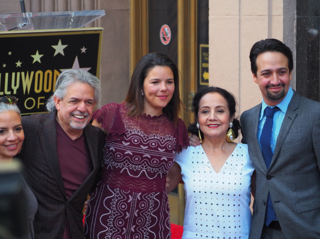 Gruppe von Menschen, die auf einem roten Teppich an der Hollywood Walk of Fame Zeremonie lächeln und posieren, mit einer Tafel, einem Glaspodest und einer Wand im Hintergrund.