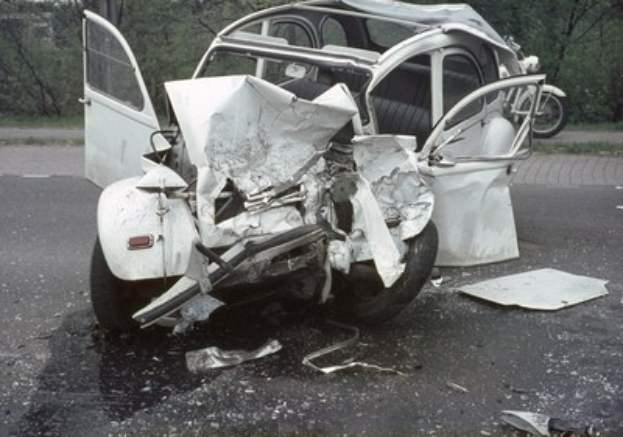 Schwerbeschädigte Limousine am Straßenrand mit Frontalaufprall, umgeben von Bäumen und einem Gebäude im Hintergrund.