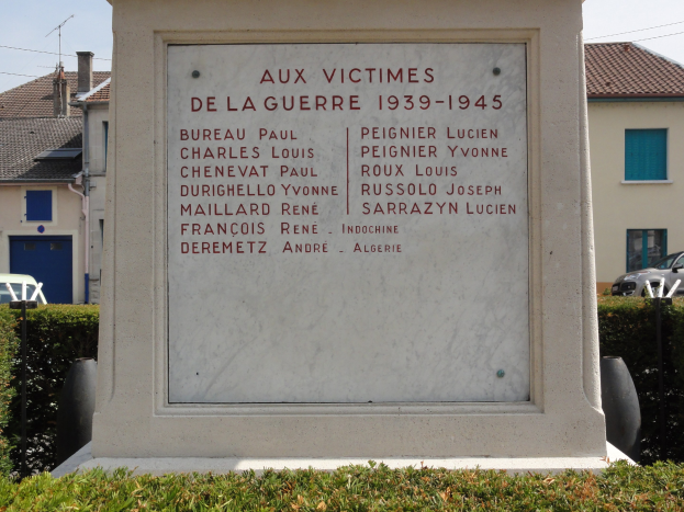 Ein Denkmal für die Opfer des Zweiten Weltkriegs (1939-1945) steht vor einem Gebäude, umgeben von Pflanzen, Fahrzeugen, Strommasten, Häusern und einem bewölkten Himmel.