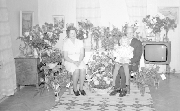 Schwarzes und weißes Foto einer Familie aus drei Personen - einem Mann, einer Frau und einem Kind - die auf einem Sofa in einem Wohnzimmer mit Blumenvasen, einem Teppich, Vorhängen und Fotorahmen im Hintergrund sitzen.