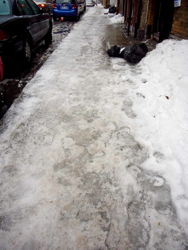 Ein von Schnee bedeckter Gehweg mit Autos oben links und einer Wand oben rechts.