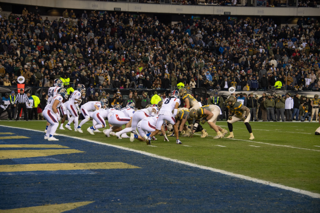 Fußballspieler in Sportkleidung und Helmen auf dem Feld während eines Spiels in einem Stadion mit Zuschauern und Anzeigetafeln im Hintergrund.