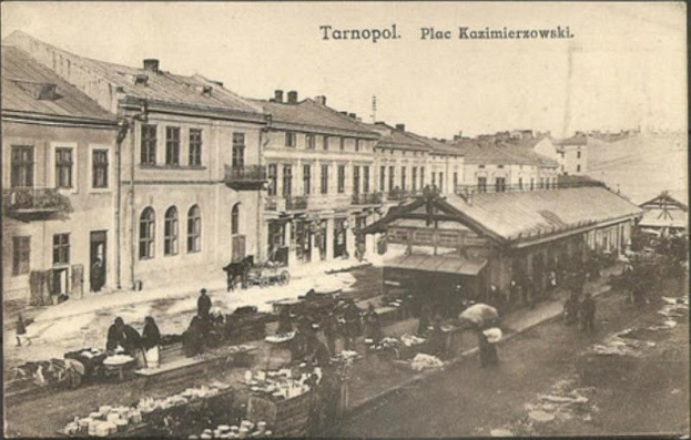 Schwarz-weiß-Foto eines belebten Marktes in Tarnopol, Polen, mit Menschen und Karren und Gebäuden im Hintergrund.