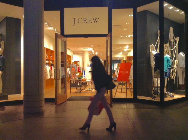 Ein Einkaufszentrum mit Glaswänden und -türen, das Schaufensterpuppen, Tische, hängende Kleider und Schränke zeigt, mit einer Frau, die daneben geht.