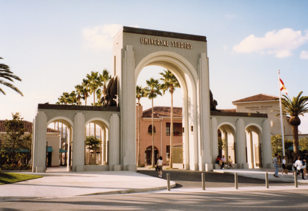 Eingang zum Universal Studios Hollywood mit einem Bogen mit Text, einer Statue, Menschen, umgeben von Grün, Gebäuden, einem Fahnenmast mit einer Flagge und einem bewölkten Himmel.
