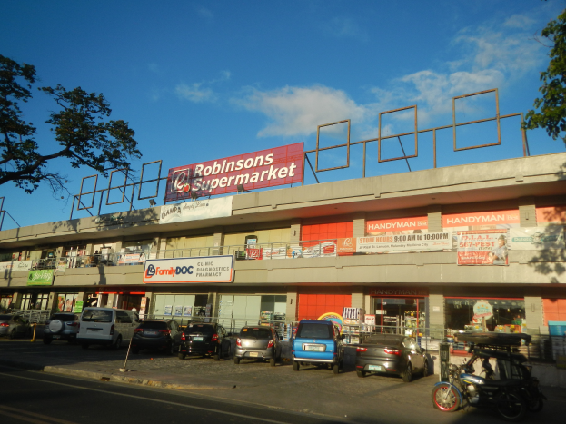 Ein Robinsons Supermarkt in Singapur mit Autos auf der Straße, einer Person auf dem Gehweg, einem Fahrrad, Pfosten, einem Schild, einem Metallrahmen, Bäumen und einem bewölkten Himmel.