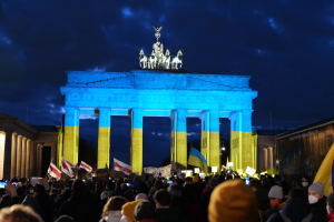Eine Menschenmenge steht vor dem Brandenburger Tor in Berlin, Deutschland, mit Fahnen und Plakaten in den Händen, einem Banner auf der rechten Seite und dem Tor mit Statuen und Säulen geschmückt unter einem bewölkten Himmel.