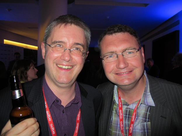 Eine Gruppe von Menschen in einem Raum, mit zwei Personen in der ersten Reihe, die eine schwarze Jacke und ein blaues kariertes Hemd tragen und in die Kamera lächeln, sowie ein Mann neben ihnen, der eine schwarze Bierflasche in der linken Hand hält und ebenfalls lächelt.