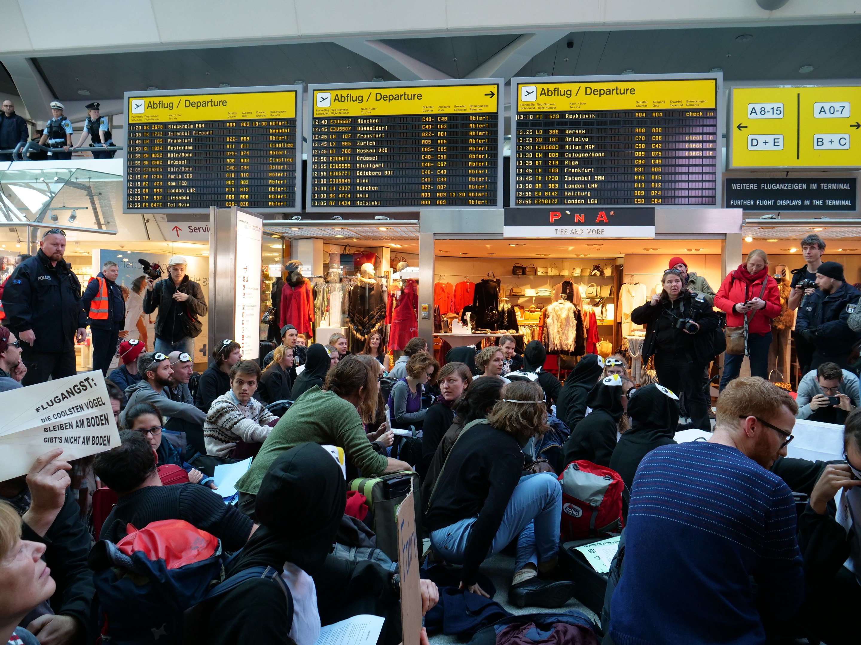 Eine große Gruppe von Menschen in einem Flughafen, einige sitzen mit Taschen und Papieren, andere stehen, mit Texttafeln und Schaufensterpuppen im Hintergrund, was auf eine Demonstration hindeutet.