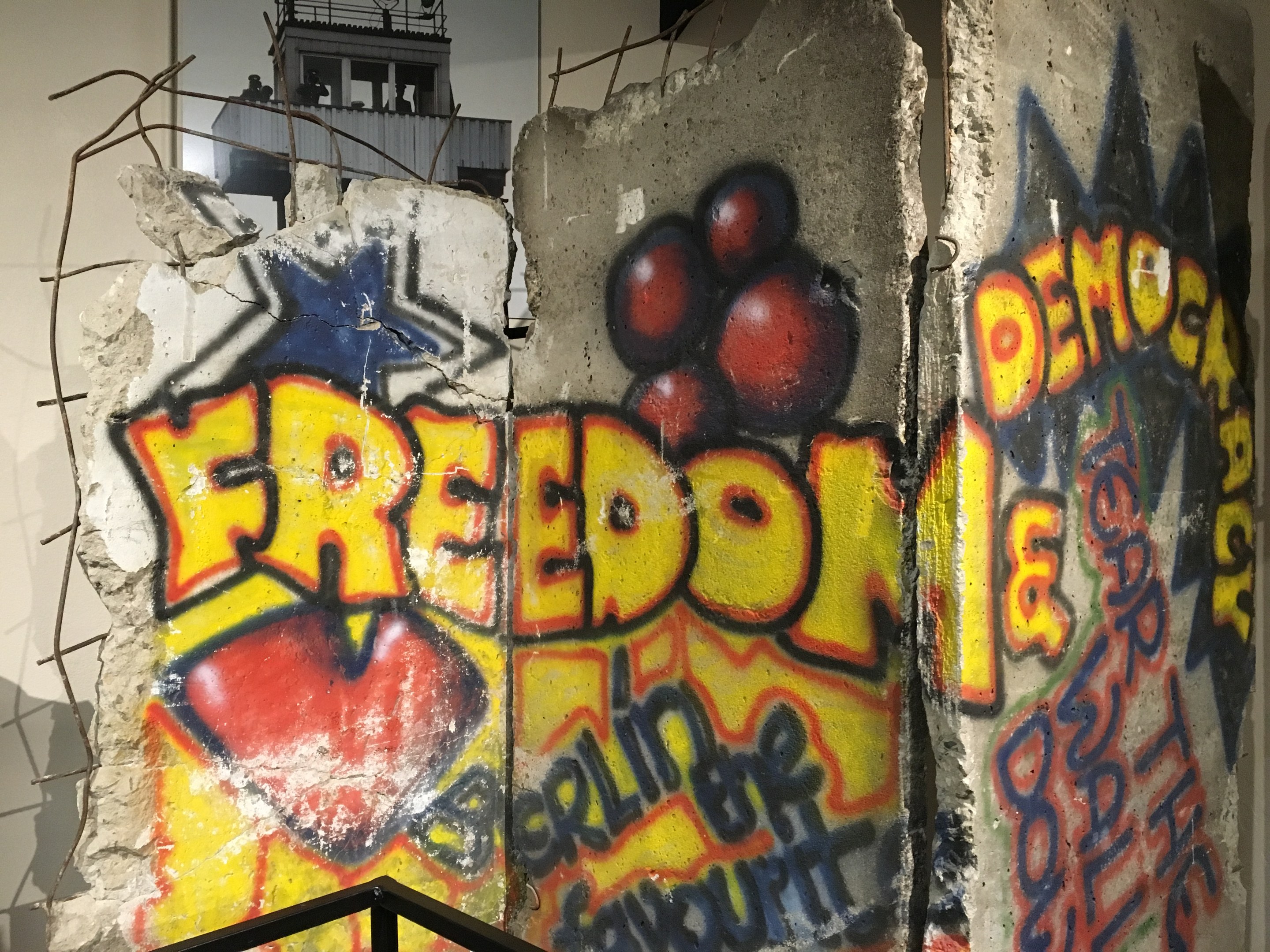 Eine Wand mit Graffiti, das in fetten schwarzen Buchstaben "Freiheit und Demokratie" lautet und unten ein Geländer sowie im Hintergrund ein Gebäude mit Fenstern zeigt.