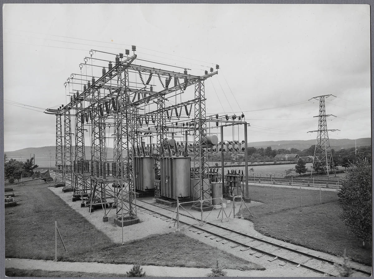 Ein altes Schwarz-Weiß-Foto einer elektrischen Umspannstation mit Transformatoren, Masten und Drähten, umgeben von Bäumen, Gras und einem Zaun, mit Eisenbahnschienen im Vordergrund und Hügeln und Himmel im Hintergrund.