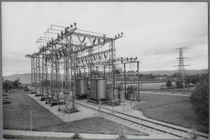 Ein altes Schwarz-Weiß-Foto einer elektrischen Umspannstation mit Transformatoren, Masten und Drähten, umgeben von Bäumen, Gras und einem Zaun, mit Eisenbahnschienen im Vordergrund und Hügeln und Himmel im Hintergrund.