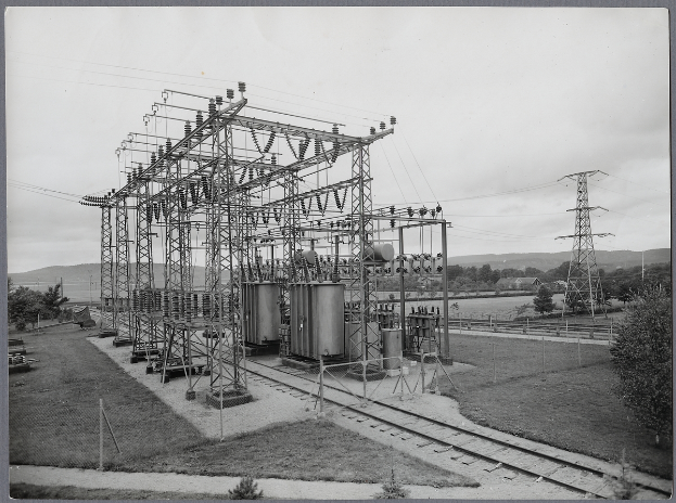 Ein altes Schwarz-Weiß-Foto einer elektrischen Umspannstation mit Transformatoren, Masten und Drähten, umgeben von Bäumen, Gras und einem Zaun, mit Eisenbahnschienen im Vordergrund und Hügeln und Himmel im Hintergrund.