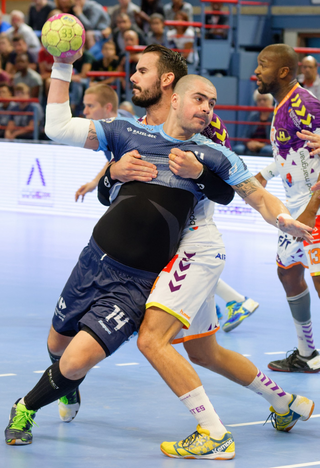 Männer spielen Handball in einer Stadionumgebung, einer hält den Ball, mit Zuschauern im Hintergrund.