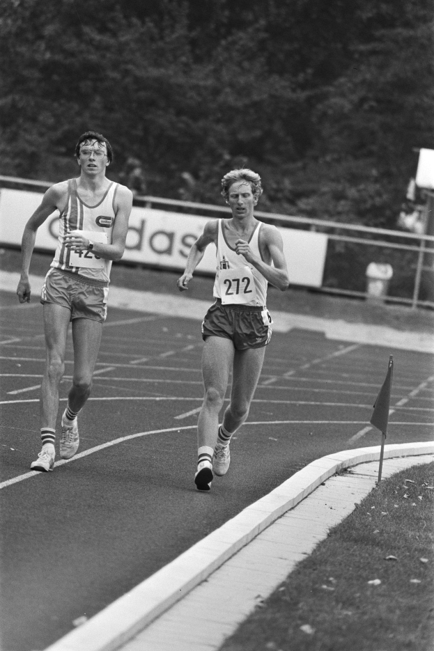 Zwei Männer laufen auf einer Laufbahn während eines Marathon, mit einem Fahnenmast auf der rechten Seite und Bäumen im Hintergrund, in Schwarz-Weiß.