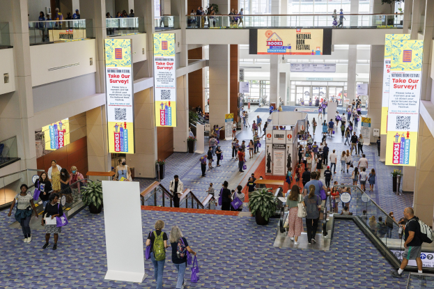 Eine große Menschenmenge bewegt sich durch ein großes Gebäude mit Bannern, Topfpflanzen, Rolltreppen, Geländern, Säulen, bedruckten Tafeln, Glasdoors und anderen Elementen, einige tragen Taschen, was auf die Teilnahme an der Nationalen Buchmesse hinweist.