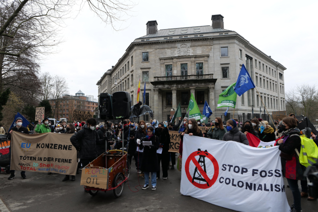 Große Gruppe von Menschen marschiert bei einer Demonstration gegen fossile Brennstoffe, trägt Transparente und Fahnen, mit einem Fahrzeug im Vordergrund und Gebäuden, Bäumen und einem klaren Himmel im Hintergrund.