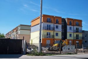 Eine Reihe von Wohngebäuden mit Fenstern, Geländern und Balkonen, einem Metallzaun, einem Pfahl, einem Bulldozer, einigen Pflanzen, einer Gruppe von Bäumen und einem bewölkten Himmel.