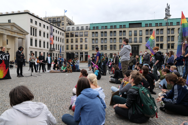Gruppe von Menschen, die auf dem Boden sitzen, vor einer Menge mit Fahnen und Transparenten bei einer Anti-Schwulen-Demonstration in Berlin, mit einer Statue, Gebäuden und einem bewölktem Himmel im Hintergrund.