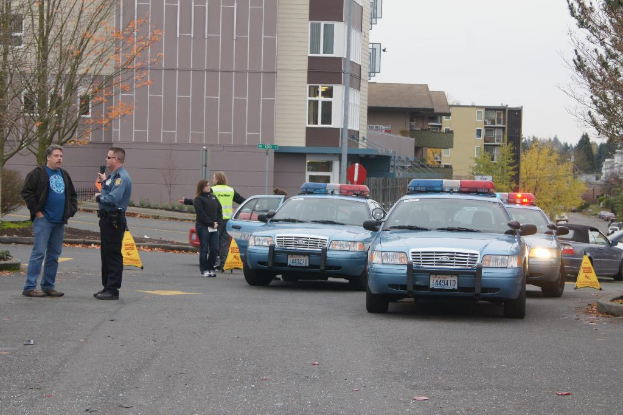 Autos auf einer Straße mit vier Personen in der Nähe, Gebäude mit Fenstern im Hintergrund, Bäume und ein Notrufsignal.