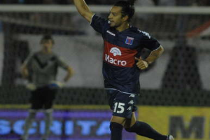 Ein Fußballer liegt auf dem Boden und feiert ein Tor, mit einer stehenden Person und einer Texttafel im Hintergrund, während eine jubelnde Menge unscharf in der Ferne zu sehen ist.