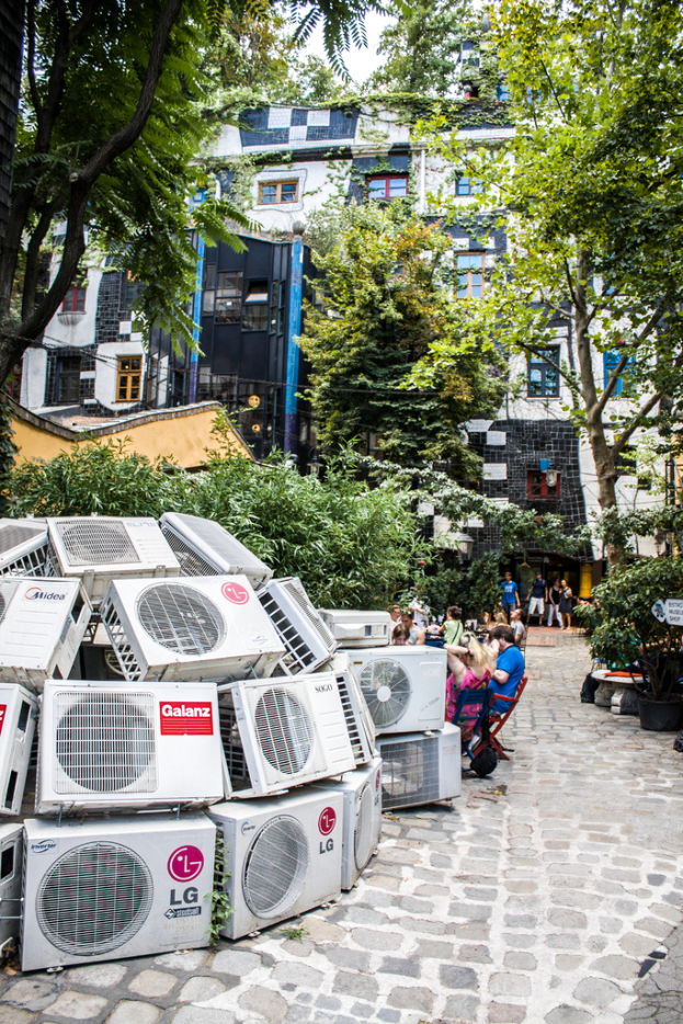 Luftkühler sind links unten auf dem Boden platziert, mit einer Straße rechts daneben, Menschen auf Stühlen sitzend und Gebäuden, Bäumen und Pflanzen oben.