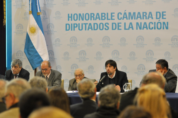 Eine Gruppe von Menschen an Tischen mit einem Krug, Papieren und einem Stift, während eine Person in der Mitte in ein Mikrofon spricht, mit einer weißen und blauen Flagge und einem Hintergrund, auf dem 'ehrenwerte kamara de' steht.