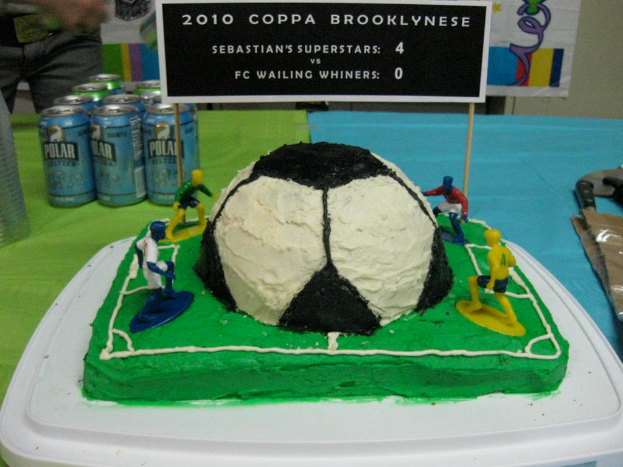 Ein fußballstadionförmiger Kuchen mit einem Fußball und vier Spielern, umgeben von Softdrink-Dosen, mit einem Banner im Hintergrund und einer Person auf der linken Seite, mit einer Tafel auf dem Kuchen.