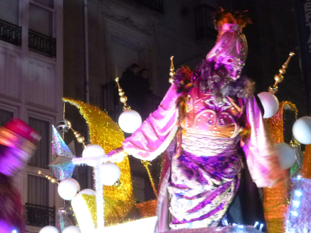 Ein Mann in einem Kostüm steht auf einem geschmückten Wagen bei einer nächtlichen Stadtparade, im Hintergrund ein Gebäude mit Fenstern und Geländern.