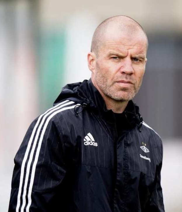 Ein kahlgeschorener Trainer in einer blauen Jacke mit weißen Streifen und einem weißen Kragen steht auf einem Fussballfeld und schaut ernst nach vorne.